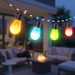 Colorful string lights hanging above a patio with people sitting around a fire pit.